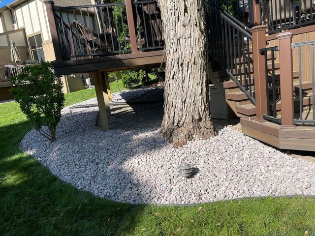Light gray stone bed with black edging encircling a large tree trunk beneath a deck and staircase.