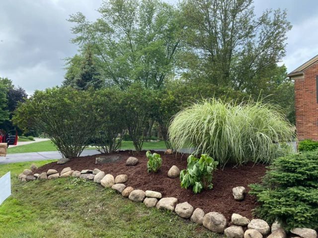 Curved mulch bed edged with fieldstone, planted with ornamental grass and shrubs along a green lawn.